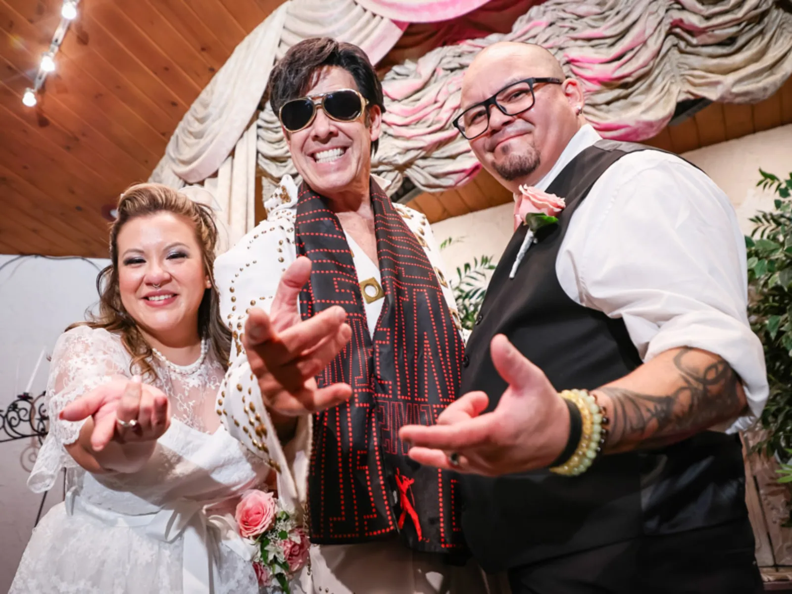 Fun Las Vegas Elvis wedding ceremony photo with smiling Elvis impersonator and joyful newlywed couple reaching toward camera inside romantic chapel venue