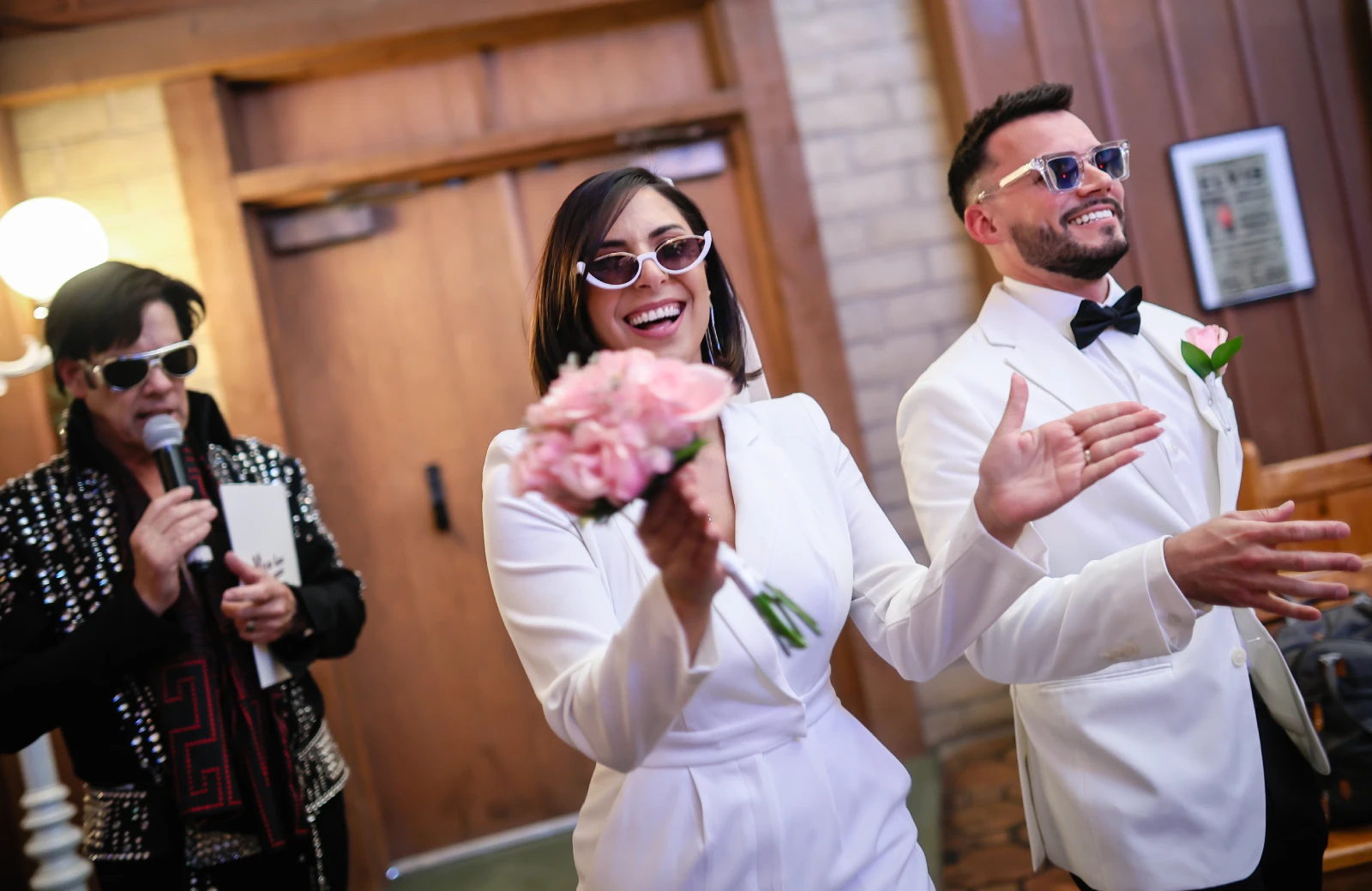 Joyful Las Vegas Elvis wedding ceremony couple in white outfits smiling and celebrating with pink bouquet while Elvis impersonator officiates inside classic chapel venue