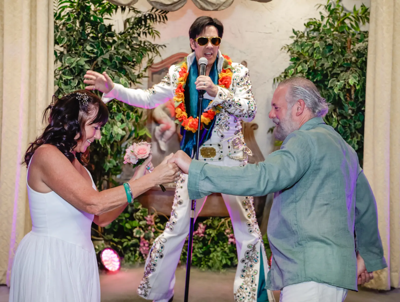 Elvis wedding ceremony moment with mature bride and groom dancing together while Elvis impersonator sings on chapel stage surrounded by greenery and soft lighting