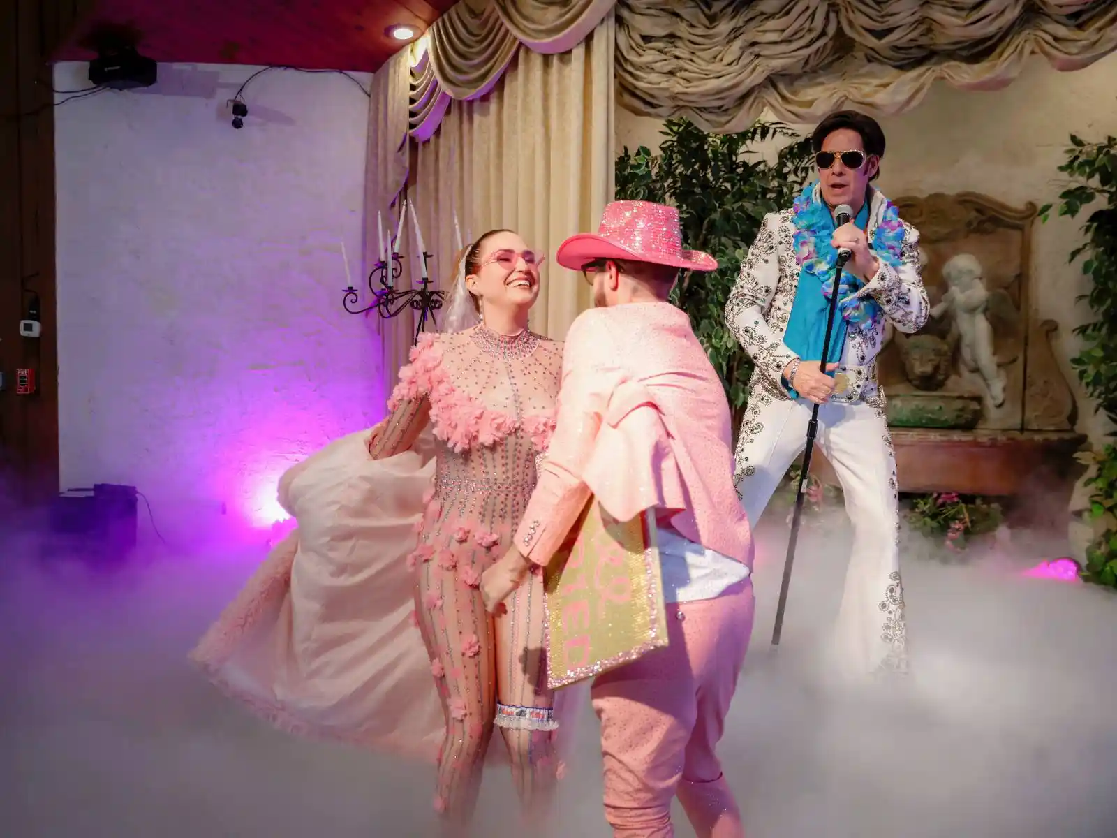 Couple in sparkly pink outfits exchanging vows with Elvis officiant and purple fog effects at a Las Vegas wedding chapel