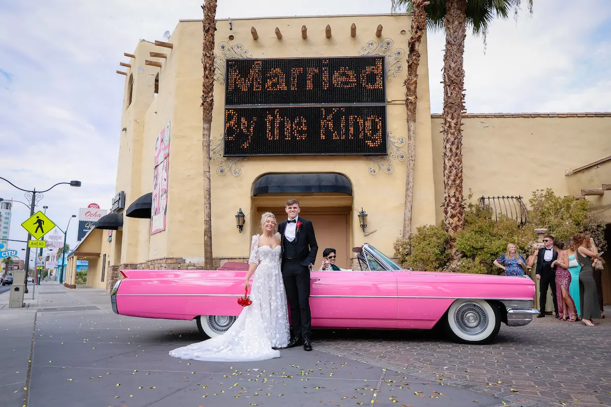 Newlyweds with pink Cadillac under Married by the King marquee sign at Viva Las Vegas Wedding Chapel