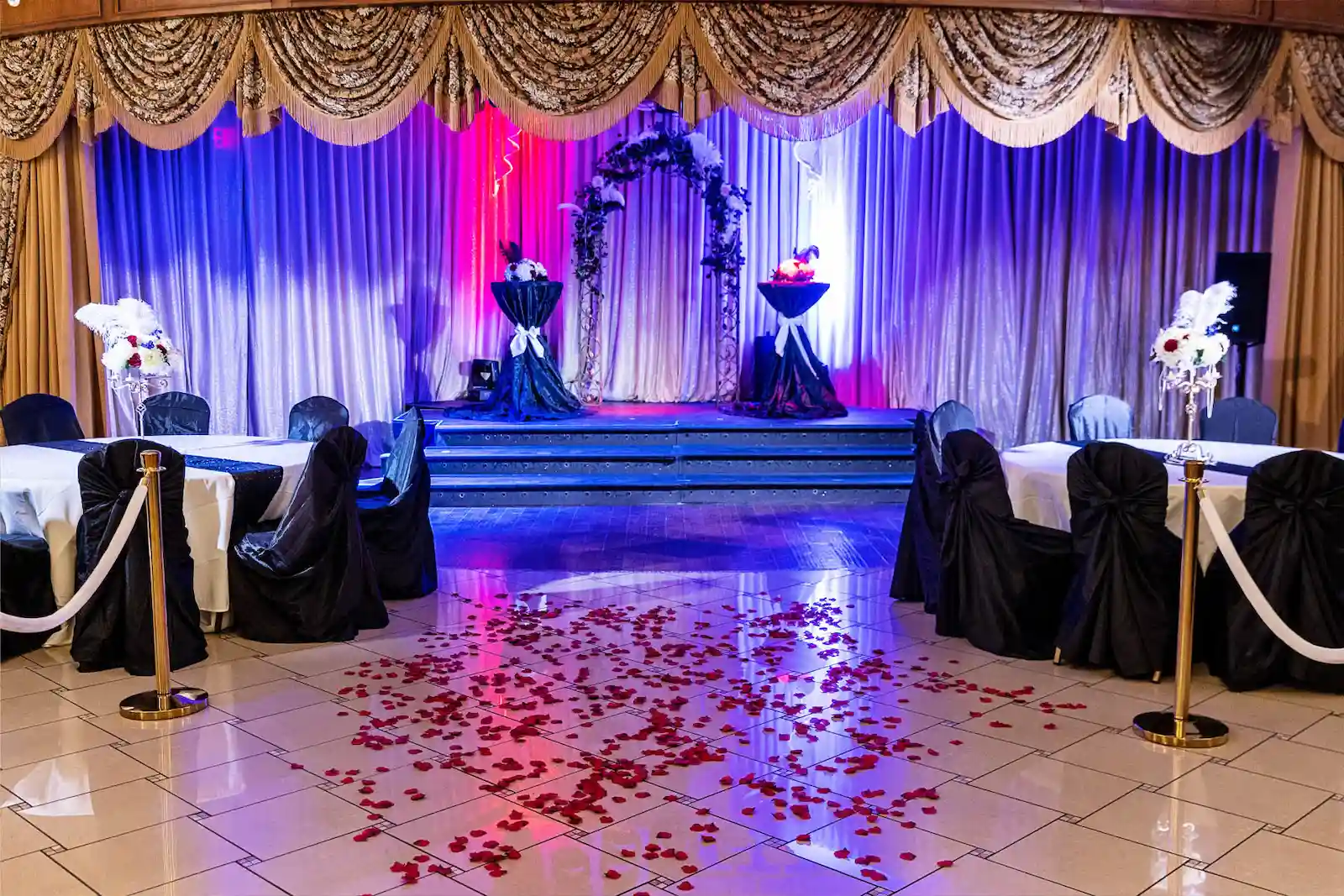 Viva Las Vegas Weddings reception hall with stage, floral arch, feather centerpieces, black chair covers, and rose petals on the dance floor