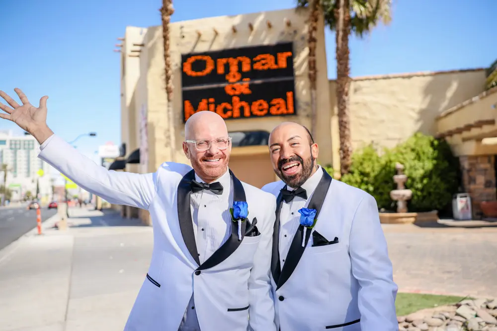 Two grooms in matching white tuxedos with blue boutonnieres celebrating in front of the Las Vegas wedding chapel marquee sign