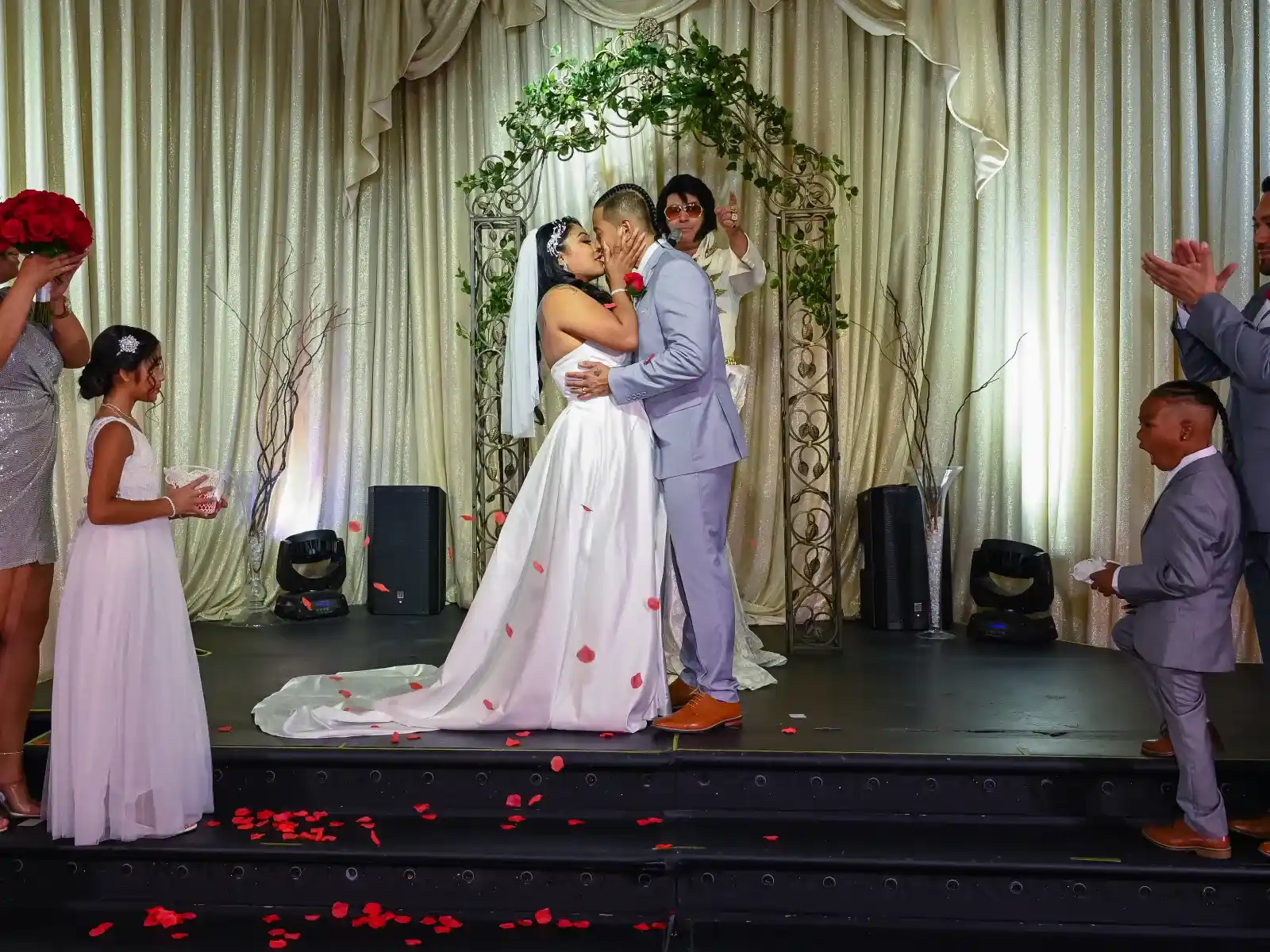 Bride and groom kissing on ceremony stage while guests throw rose petals during wedding celebration.
