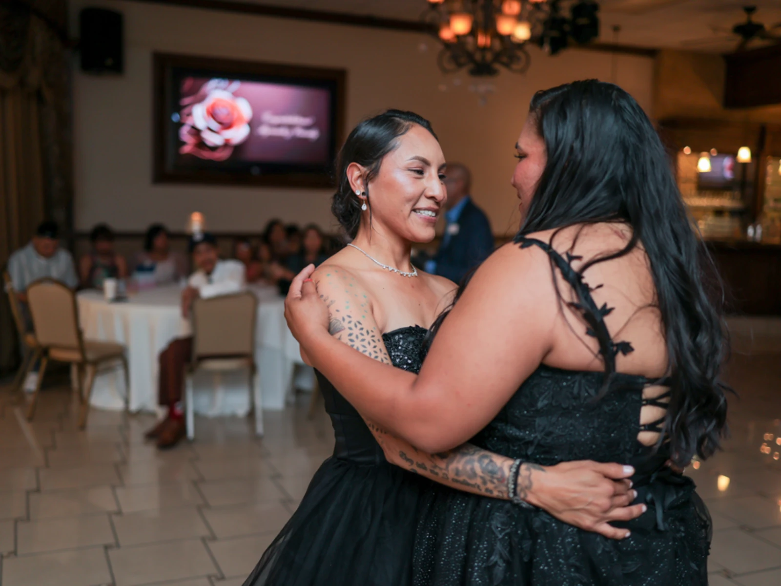Same-sex wedding couple dancing during reception in elegant ballroom with guests in background.
