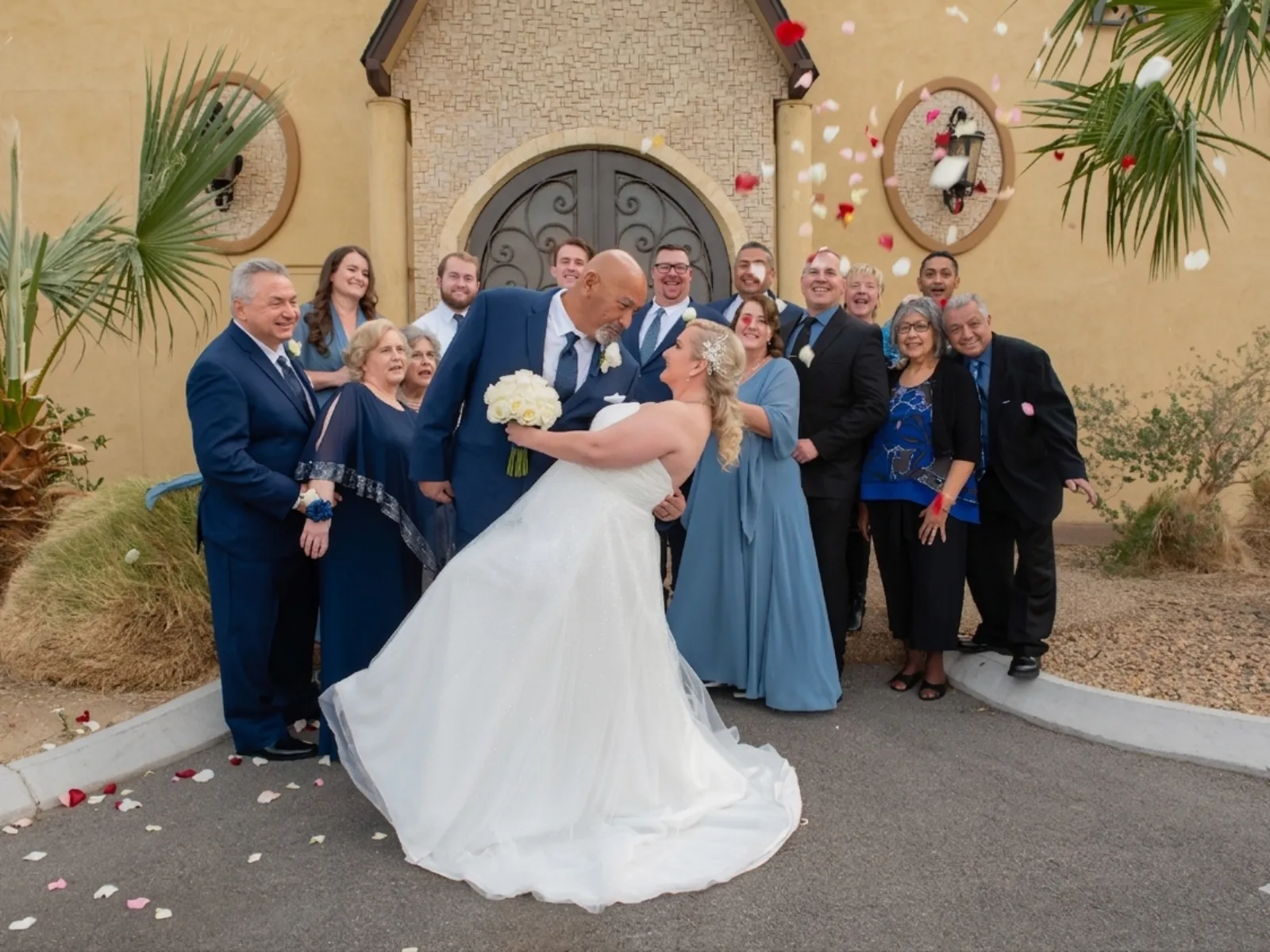 Bride dipped by groom in front of chapel surrounded by joyful family wedding group outdoors.