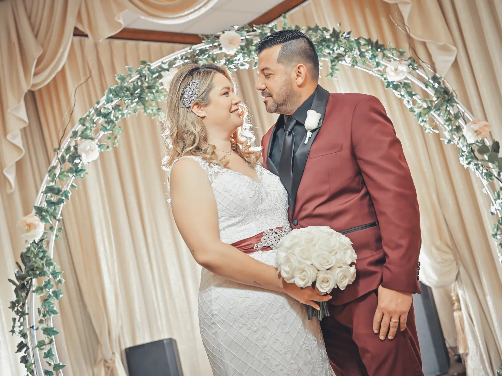 Newlywed couple posing under floral ceremony arch indoors with burgundy groom suit and white bouquet.