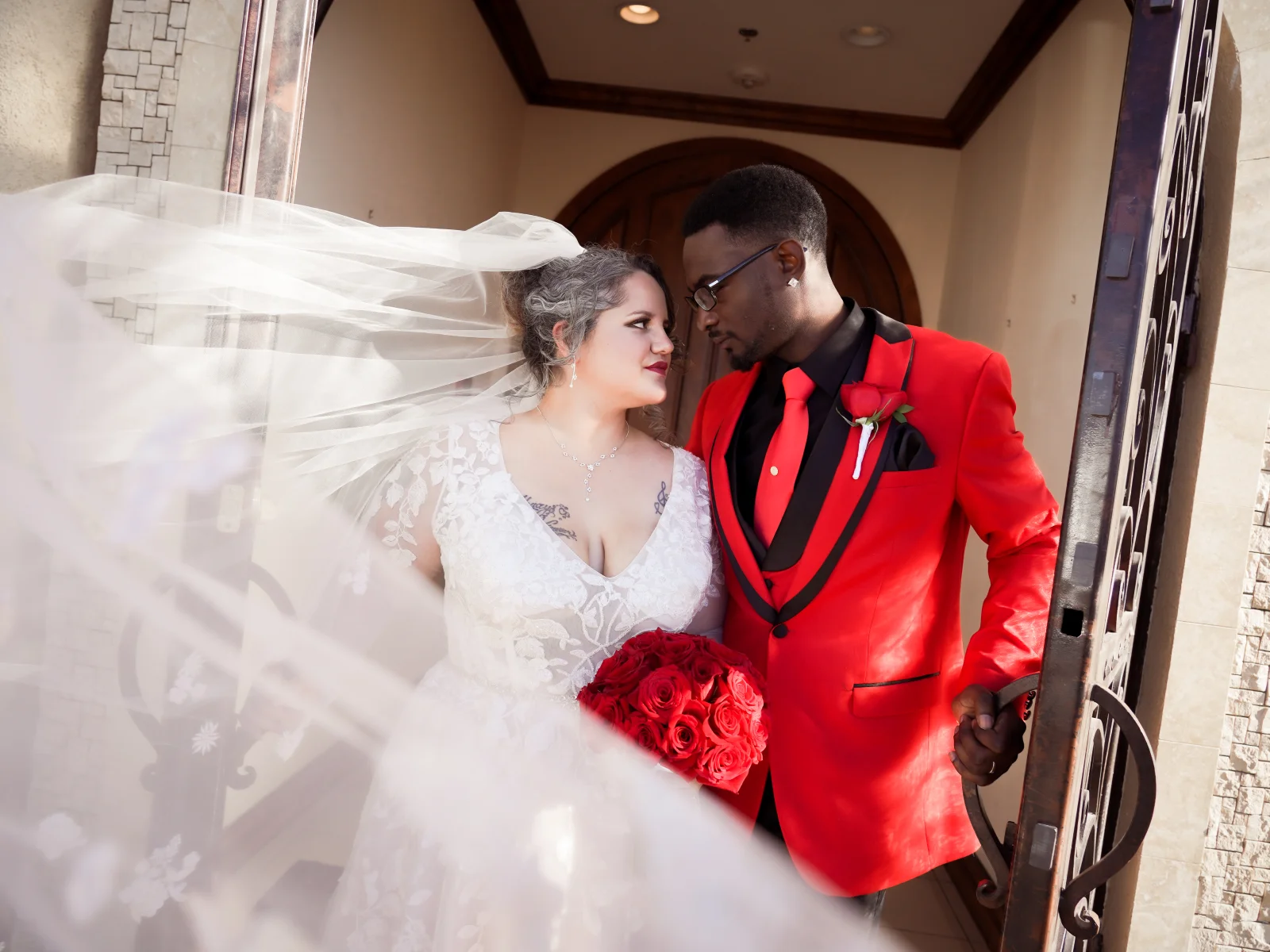 Wedding couple standing in doorway with flowing veil and groom in red suit.