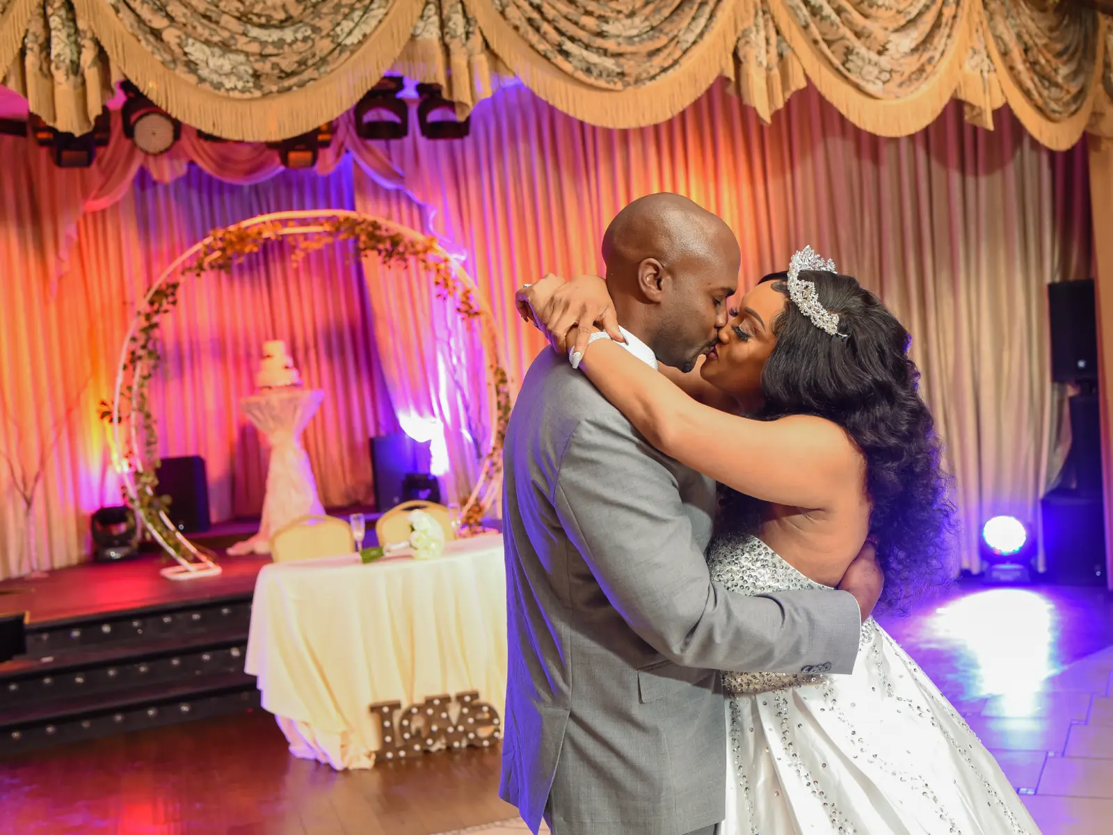 Newlywed couple kissing on dance floor with colorful wedding lighting and decor.