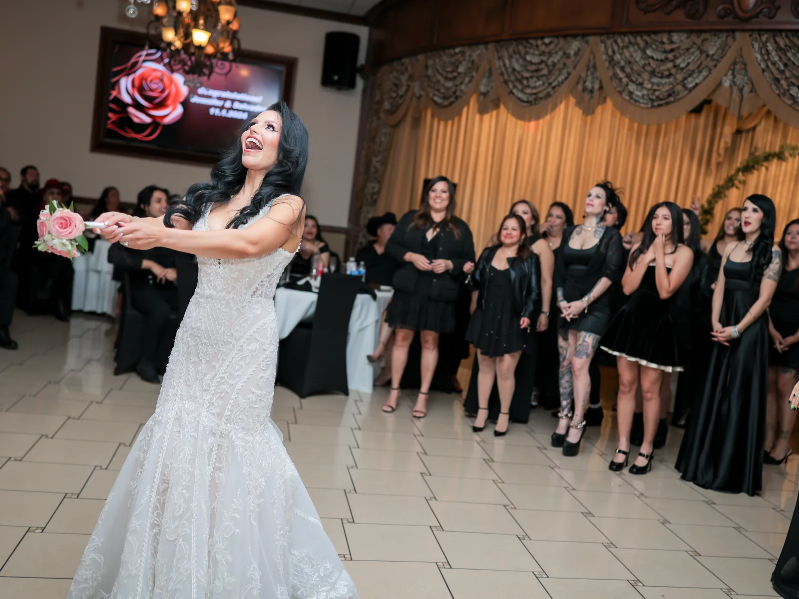 Bride tossing bouquet toward excited guests dressed in black at wedding reception.