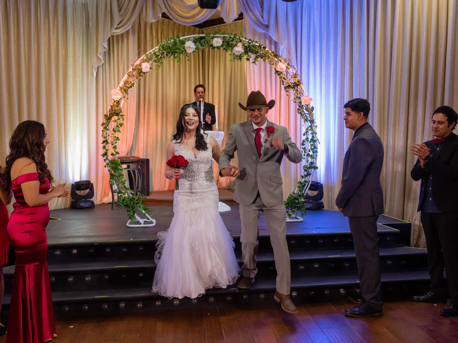 Bride and groom walking down stage steps under floral circle arch after ceremony.