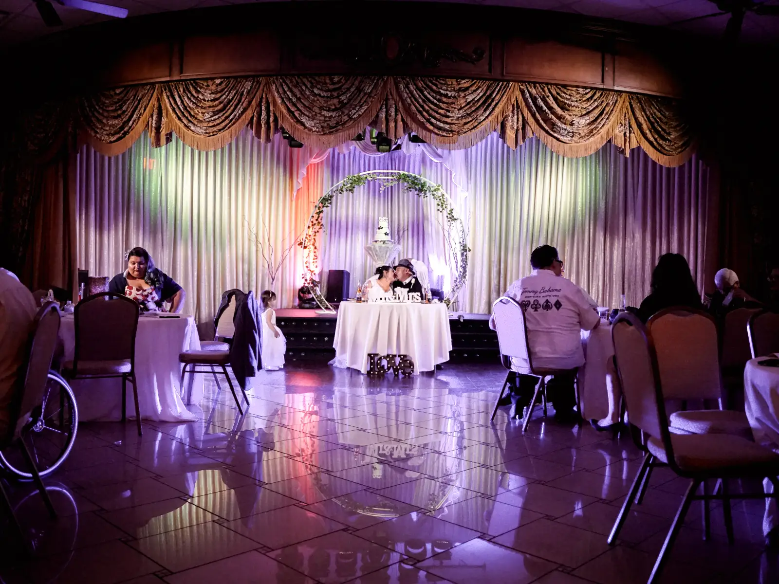 Newlyweds kissing at sweetheart table during colorful lit reception in banquet hall.