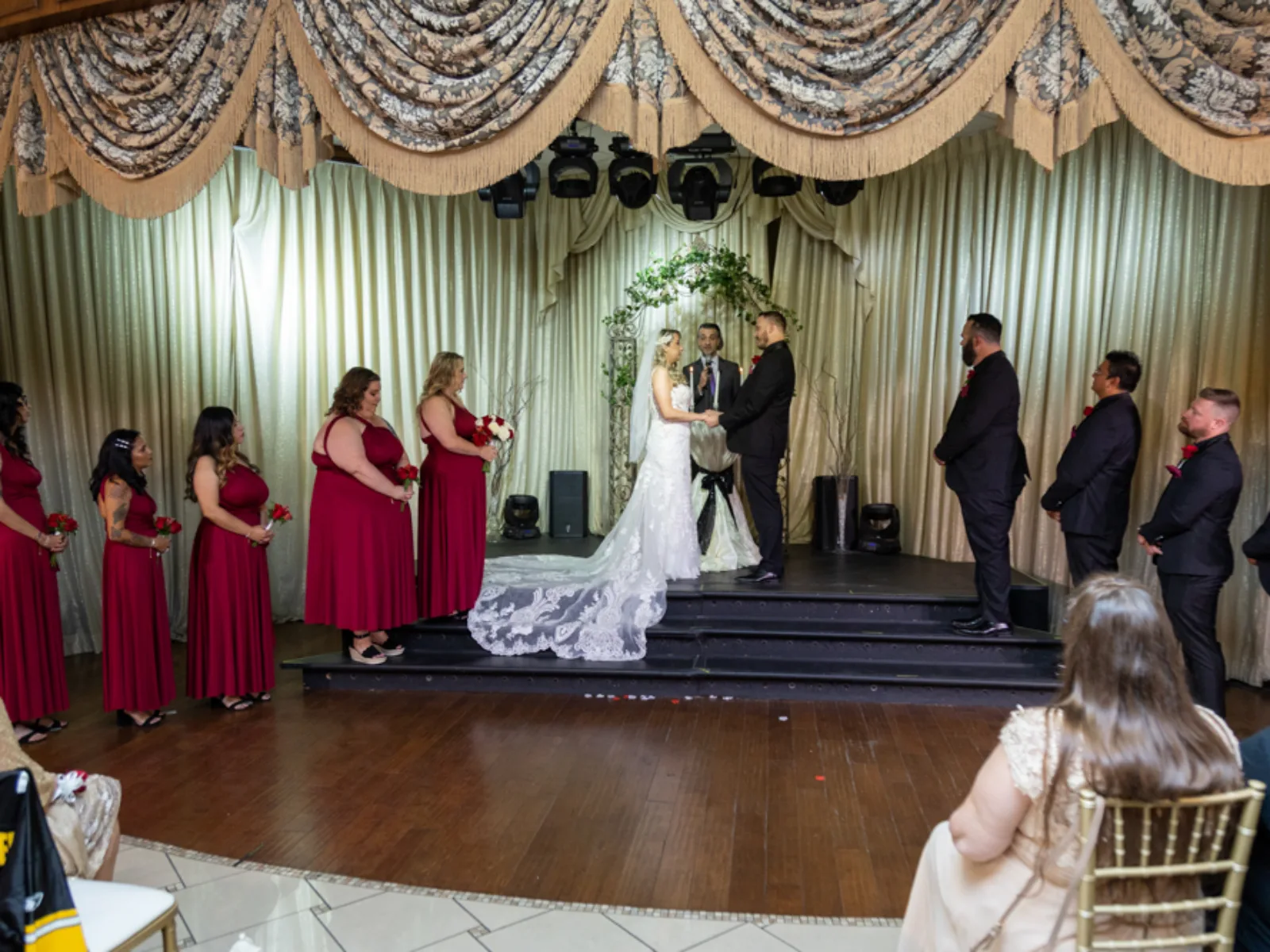 Couple exchanging vows on small stage with red-dressed bridesmaids and groomsmen lined up.