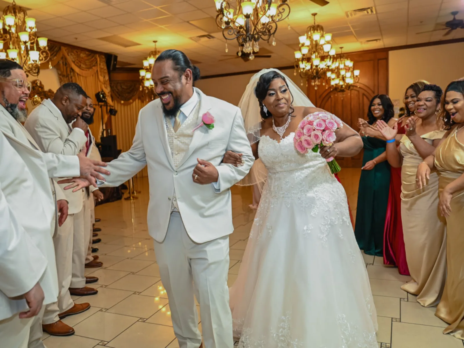 Bride and groom in white outfits celebrating grand entrance with wedding party clapping indoors.
