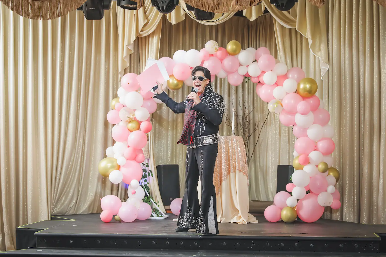 Elvis impersonator performing on stage under a pink and gold balloon arch at a Las Vegas wedding reception