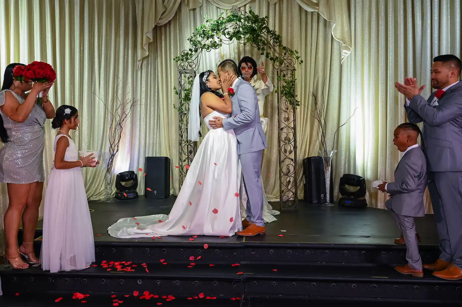 Bride and groom sharing first kiss on stage with rose petals and Elvis officiant at a Las Vegas wedding reception